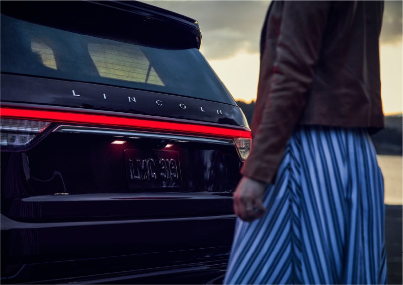 A person is shown near the rear of a 2023 Lincoln Aviator® SUV as the Lincoln Embrace illuminates the rear lights | Doggett Lincoln of Beaumont in Beaumont TX